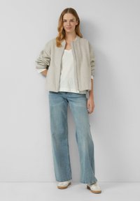Lichtgrijze bomberjack met geribbelde boorden, rits aan de voorkant, gecombineerd met een witte V-hals shirt en brede blauwe jeans; model staat in een neutrale omgeving.
