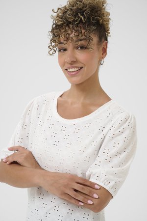 Blouse blanche avec un design texturé et des motifs à œillets, un col rond et des manches courtes puff. Tissu lisse et coupe décontractée.