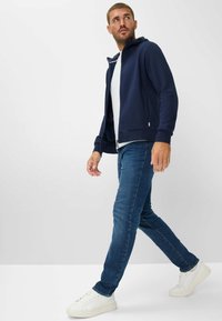 Dunkelblaues Zip-Hoodie mit einer Vorderentasche, kombiniert mit eng anliegenden dunkelblauen Jeans und weißen Sneakern, vor einem schlichten hellen Hintergrund.