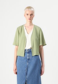Lys olivenfarget croppet topp med korte ermer, åpen frontdesign, båret over en hvit V-hals skjorte og et blått denim skjørt med en frontsplitt.