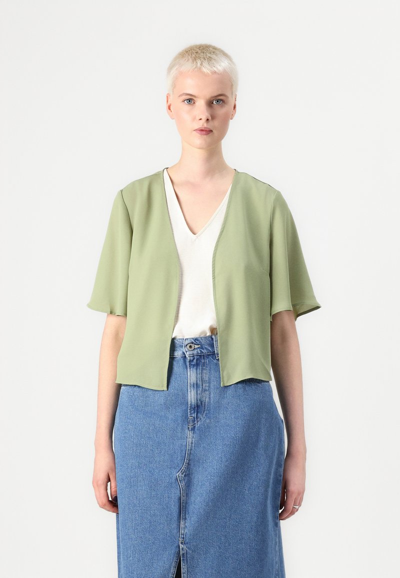 Lys olivenfarget croppet topp med korte ermer, åpen frontdesign, båret over en hvit V-hals skjorte og et blått denim skjørt med en frontsplitt.