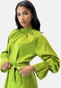 Hellegrüne Satinbluse mit hohem Kragen, puffartigen Ärmeln und einer gebundenen Taille. Glatte Textur mit leichtem Glanz.
