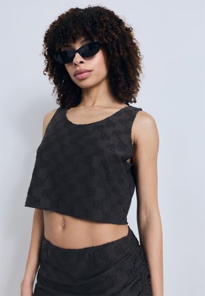 Femeie cu păr creț purtând un top fără mâneci negru texturat și o fustă drapantă, ochelari de soare negri cat-eye, pozând pe un fundal simplu.