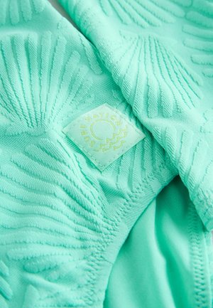 Pūsta žalių mėtos atspalvio tekstūruota medžiaga su iškilusiais kriauklių raštais, viename kampe siūtas geltonas etiketės užrašas „SURF NATION".