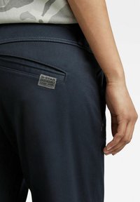 Donkerblauwe broek met een gestructureerde stof, zichtbare stiksels en een rechthoekige merkpatch aan de achterkant. Ook voorzien van zijzakken.