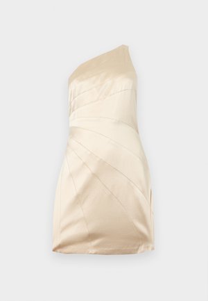 Pikkade tumedate juustega naine, kes kannab ühe õlaga heledat beežikat satiinist minikleiti ja kuldseid kõrgete kontsadega sandaale, seisab tavalise tausta ees.