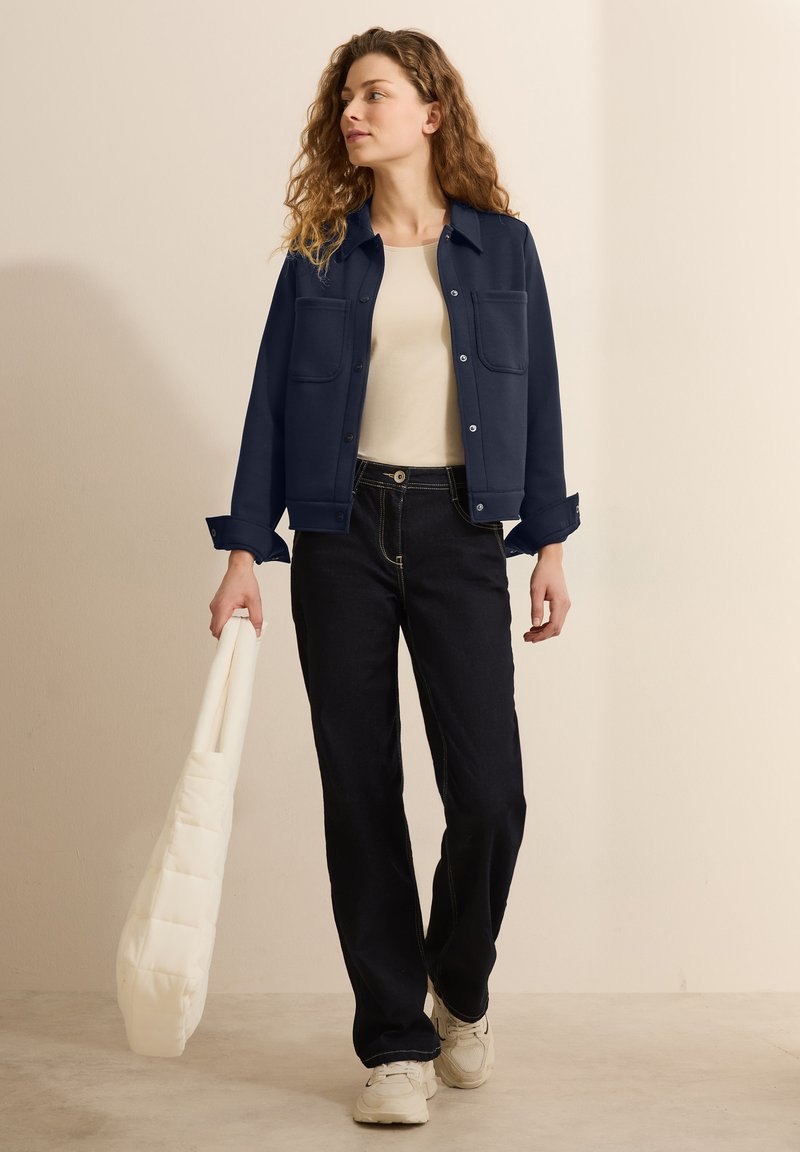 Námořnická modrá overshirt s dvěma předními kapsami, stylizovaná přes béžový top, spárovaná s tmavými džíny a bílými teniskami, držící krémovou kabelku.