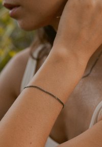 Thin metallic chain bracelet in dark gray, featuring a simple interlocking design; worn on a tanned wrist. Smooth texture, minimalistic style.