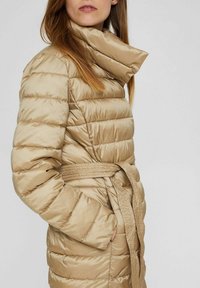 Beige gepolsterter Mantel mit hohem Kragen, tailliertem Gürtel und gestepptem Design. Glatte Textur mit sichtbaren horizontalen Nähten.