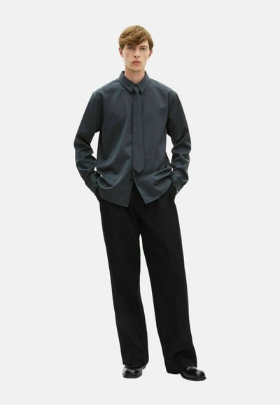 Dunkelgrauhe Hemd mit Kragen, kombiniert mit schwarzen weiten Hosen und schwarzen Schuhen. Der Stoff wirkt strukturiert und texturiert.