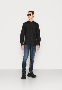 Camicia nera a maniche lunghe con bottoni, abbinata a jeans blu sbiaditi e sneakers nere. Il modello indossa occhiali da sole rotondi neri.
