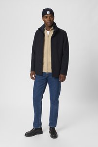 Cappotto in misto lana blu navy con colletto rialzato, sovrapposizione beige con zip, jeans in denim blu e scarpe slip-on nere. Berretto in maglia di cotone.