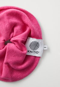 Roze gebreide scrunchie van zacht materiaal met een cirkelvorm. Heeft aan de ene zijde een wit label met zwart logo en tekst.