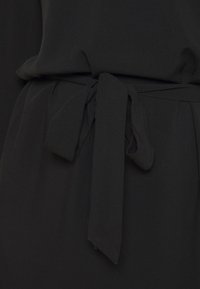 Černý kravata pás vyrobený z jemné tkaniny, elegantně uvázaný v pase. Šaty mají hladkou texturu a čistý, jednoduchý design.