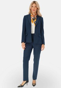 Granatowy garnitur dopasowany, z dwoma guzikami, bocznymi kieszeniami i dopasowanymi spodniami; zestawiony z białym topem i kolorowym szalikiem. Czarny obcas dopełnia stylizację.