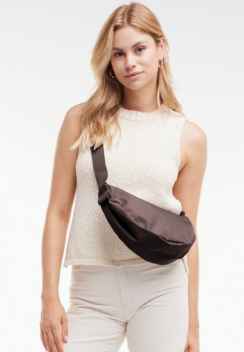 Blonde Frau mit ärmellosem beigen Stricktop und weißer Hose, die eine schwarze Umhängetasche mit verstellbarem Riemen trägt.