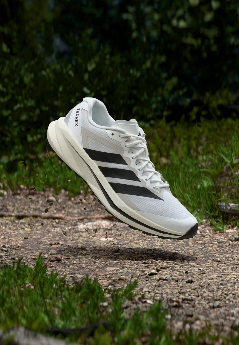 Λευκό παπούτσι τρεξίματος Adidas Terrex με μαύρες ρίγες, αιωρούμενο πάνω από ένα χαλίκι μονοπάτι περιτριγυρισμένο από πράσινη βλάστηση.