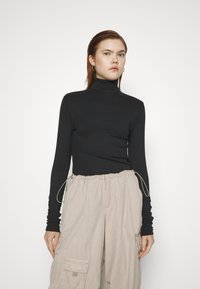Svart tettsittende genser med høy hals laget av rillestoff, kombinert med beige cargobukser med lommer og snor i livet.