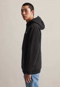 Zwarte capuchontrui met een losse pasvorm, geribbelde manchetten en een voorkantel. Gecombineerd met lichtblauwe jeans, wat een casual uitstraling geeft.