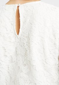 Hvid blondetop med blomster mønstre, en nøglehulslukning bagpå og struktureret stof. Designet fremviser detaljeret håndværk og bløde tråde.