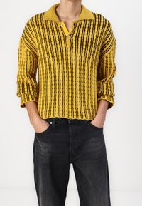 Żółto-czarny sweter w paski z kołnierzykiem i listwą na guziki. Noszony na ciemnych dżinsach, prezentuje luźny krój i dzianinową fakturę.