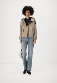 Giacca cropped beige con cappuccio, chiusura a bottoni, tasche laterali, indossata sopra una camicia blu navy, abbinata a jeans azzurri chiari e stivali neri.