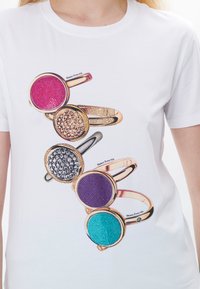 Camiseta blanca con una ilustración de cuatro anillos con piedras coloridas: rosa, púrpura, gris y turquesa, en bandas de oro y plata.