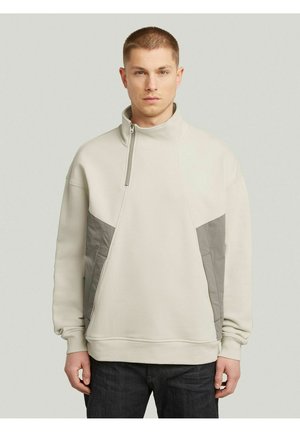 Homme portant un pull beige clair à col montant et des panneaux gris sur les côtés, debout devant un fond uni gris clair.