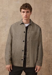 Camicia a quadri in tessuto marrone e beige, con colletto, bottoni frontali e maniche lunghe. Indossata sopra un maglione scuro.