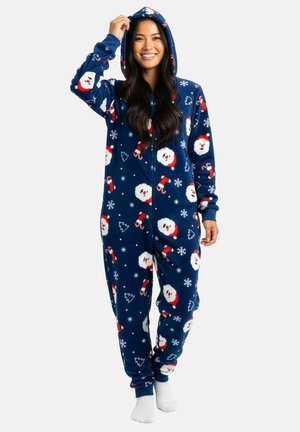 Frau lächelt und trägt einen blauen Kapuzen-Overall mit Santa-Gesichtern, Zuckerstangen, Schneeflocken und Weihnachtsbaum-Mustern, steht barfuß.