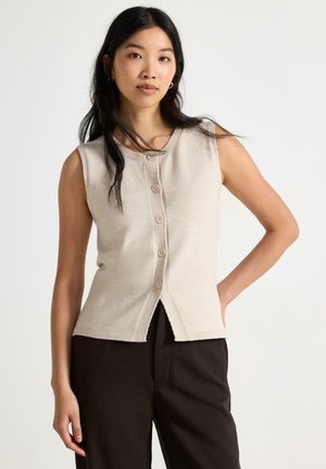 Donna con lunghi capelli scuri che indossa un gilet beige senza maniche abbottonato e pantaloni neri, in piedi con una mano sui fianchi.