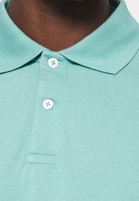 Petrolejová polo košile vyrobená z bavlny s texturovaným povrchem. Obsahuje klasický límec, tříknoflíkovou légu a malou přední kapsu.