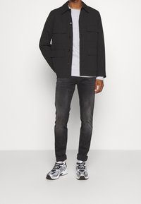 Svart knappad jacka med flera fickor fram, bärs över en ljusgrå sweatshirt, i kombination med mörkgrå jeans och silverfärgade träningsskor.