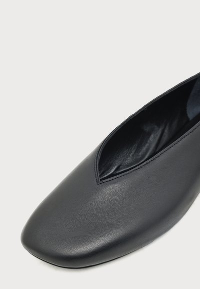 Gros plan d'une chaussure plate en cuir noir avec un bout carré arrondi et une surface supérieure lisse sur un fond clair.