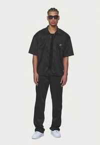 Camisa preta de manga curta com botões e calças combinando, feitas de tecido liso. Apresenta um bolso frontal e tênis brancos. Estilizado com óculos de sol.