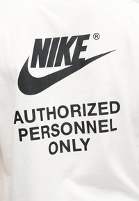 Λογότυπο Nike πάνω από έντονο κείμενο που αναγράφει "ΜΟΝΟ ΕΞΟΥΣΙΟΔΟΤΗΜΕΝΟ ΠΡΟΣΩΠΙΚΟ" σε λευκό υφασμάτινο φόντο, πιθανώς μέρος ενδύματος.