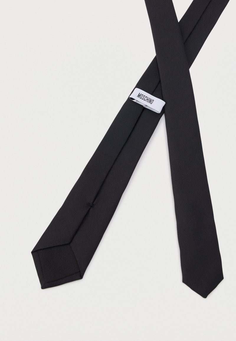 Cravate noire fine étalée à plat montrant l'avant et l'arrière, avec une étiquette en tissu blanc portant l'inscription "MOSCHINO" au dos de la cravate.