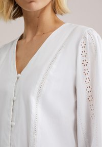 Blouse blanche avec col en V, manches longues détaillées et boutons en tissu. Les accents d'épaule bordés de dentelle ajoutent texture et intérêt.
