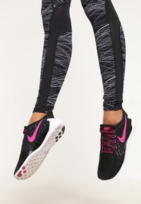 Černé sportovní boty s růžovými akcenty, s texturovaným síťovaným svrškem a bílou gumovou podešví. Nosené s černými legínami s potiskem.