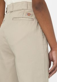 Detailní záběr na béžové pracovní kalhoty s kapsou na zadní straně a štítkem značky Dickies na pasu, které má na sobě osoba s tmavou pletí.