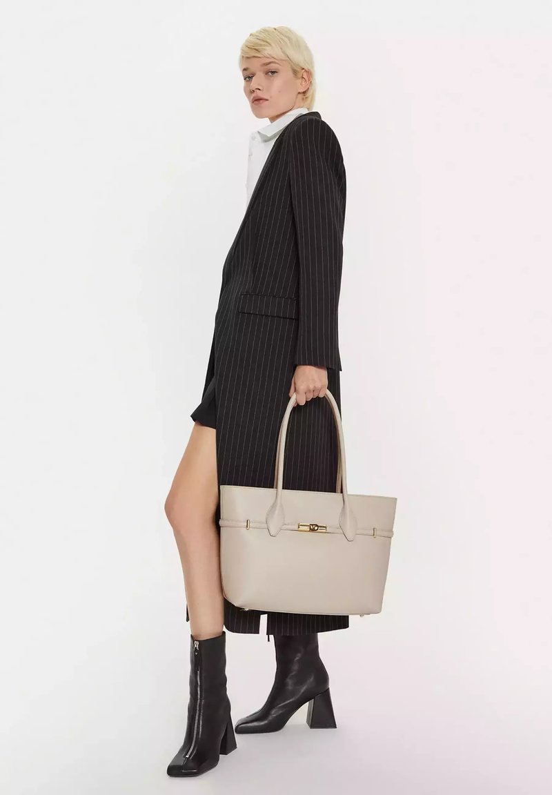 Beige leren tote bag met twee bovenhandvaten, plat ontwerp, goudkleurige hardware en zichtbare stiksels. Model draagt een gestreepte blazer en zwarte laarzen.