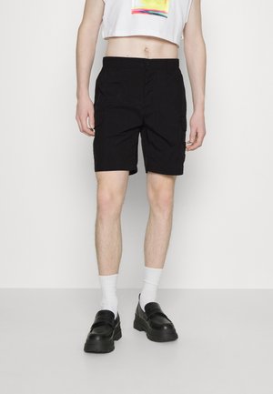 Schwarze Cargo-Shorts mit lockerer Passform, ausgestattet mit Seitentaschen und einer glatten Textur. Kombiniert mit weißen Crew-Socken und klobigen schwarzen Schuhen.