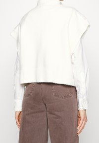 Weißer, gestrickter ärmelloser Pullover mit geripptem Kragen und Saum, getragen über einem weißen Hemd mit Kragen, kombiniert mit braunen, weit geschnittenen Hosen.