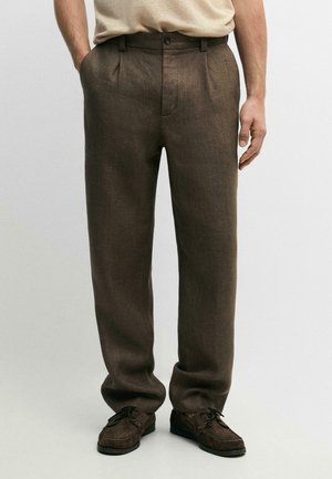 Mann trägt dunkelbraune gerade geschnittene Hose und dunkelbraune Schnürschuhe, steht mit teilweise in den Taschen steckenden Händen.