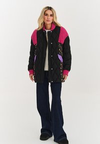 Čierna prešívaná bunda s ružovými pletenými ramenami, kvetinovými bočnými akcentmi, fialovými vreckami a predným zapínaním na cvoky. Nosená s tmavými rifľami.