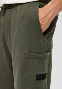 Grüne Baumwoll-Sweatpants mit strukturiertem Finish, ausgestattet mit Seitentaschen und einer Cargo-Tasche, akzentuiert durch ein schwarzes Markenpatch.