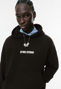 Ung person med fletninger iført en sort hættetrøje med teksten "STWD STUDIO", en blå stribet skjortekrave og en sølvvedhængs halskæde.