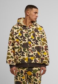 Felpa con zip in tessuto mimetico nei toni del beige, verde e marrone, decorata con faccine sorridenti gialle e dotata di tasca frontale. Etichetta del marchio sull'orlo.