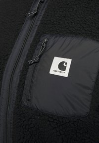 Mustapohjainen fleece-takki, jossa on sileä musta nailon-tasku. Varustettu vetoketjukiinnityksellä ja valkoisella neliölogo-patchilla, jossa on musta graafinen design.