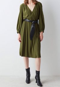 Femme portant une robe midi vert olive à manches longues et col en V profond, ceinture noire nouée à la taille, et bottines noires à talons moyens.